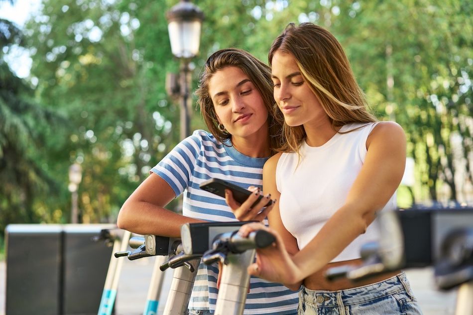 Two women looking at a phone riding scooters